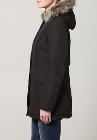 Černá parka s kapucí podšitou kožešinou, předními kapsami a zapínáním na zip/tlačítka. Má texturovanou látku a mírně přiléhavý střih.