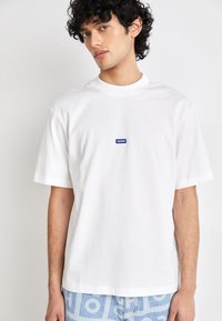White T-shirt in cotton with a crew neck. Features a blue rectangular logo patch reading "HUGO" on the chest. Short sleeves, relaxed fit.
