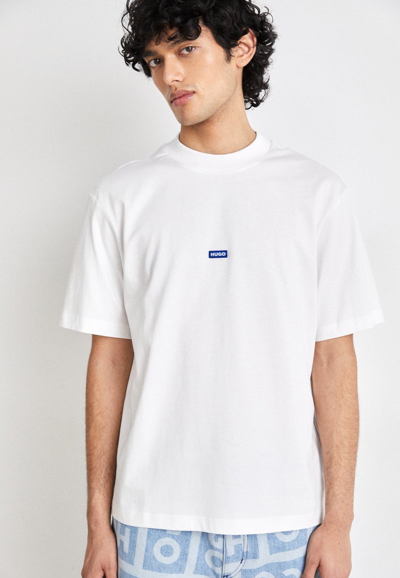 White T-shirt in cotton with a crew neck. Features a blue rectangular logo patch reading "HUGO" on the chest. Short sleeves, relaxed fit.
