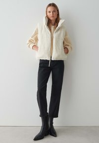 Witte, fluffy mouwloze vest met een hoge kraag, gecombineerd met een lichte trui, zwarte tapering broek en zwarte puntige enkelboots.
