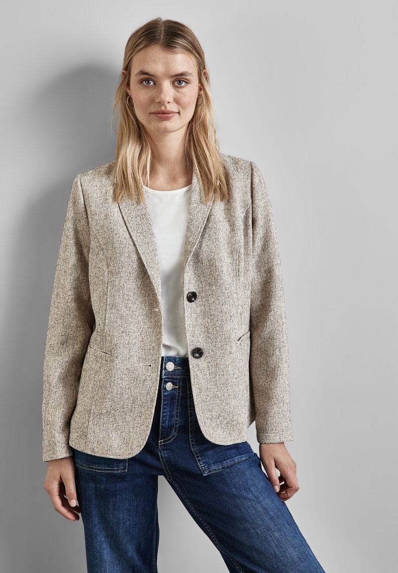 Beige strukturierter Blazer mit taillierter Form, Reverskragen, zwei schwarzen Knöpfen und Fronttaschen, kombiniert mit einem weißen Oberteil und dunklen Jeans.