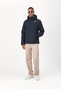 Chaqueta impermeable azul marino con capucha, cierre de cremallera y logo en el pecho, combinada con pantalones beige y zapatillas blancas.