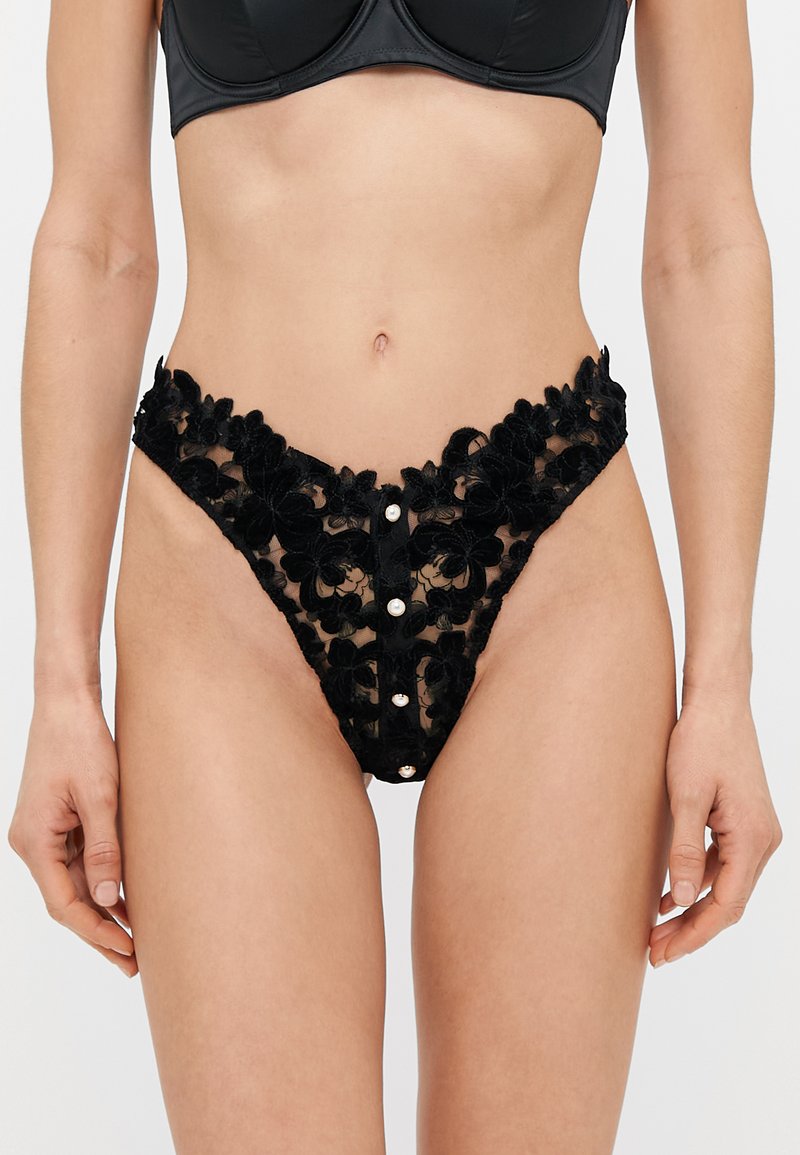 Femme portant des sous-vêtements en dentelle florale noire avec des boutons en perle à l'avant et un soutien-gorge noir assorti, montrée de la taille aux cuisses.