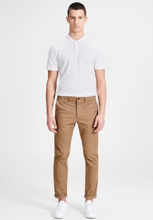 Chemise polo blanche à manches courtes, associée à un pantalon chino marron et des baskets blanches. La tenue présente une coupe classique avec un design simple.