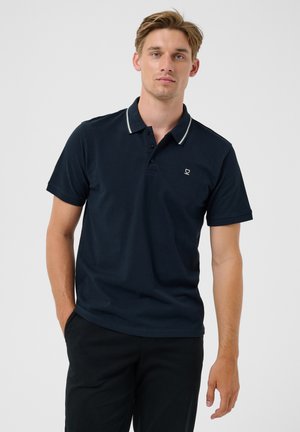 Joven con pelo claro que lleva una camiseta polo azul marino oscura con ribete blanco en el cuello y pantalones negros, de pie frente a un fondo claro y liso.