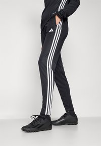 Schwarze Sporthosen mit weißen Seitenstreifen, taillierter Schnitt und mit Reißverschlüssen versehenen Taschen. Kombiniert mit schwarzen Sportschuhen mit strukturierten Sohlen.