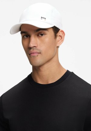 Homme portant une casquette blanche avec le logo « BOSS » et un t-shirt noir col rond, face à un fond clair uni.