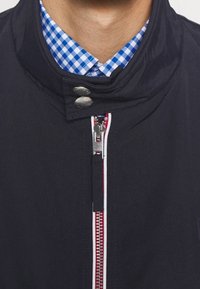 Granatowa kurtka z wysokim kołnierzem, srebrnymi zatrzaskami, czerwono-białym detalem zamka błyskawicznego oraz niebieską koszulą w kratę pod spodem.