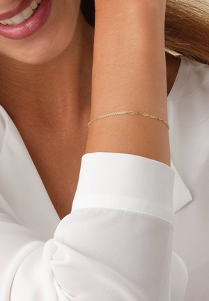 Lächelnde Person, die ein zartes goldfarbenes Armband am Handgelenk und eine weiße Bluse mit langen Ärmeln trägt, deren Manschette sichtbar ist.