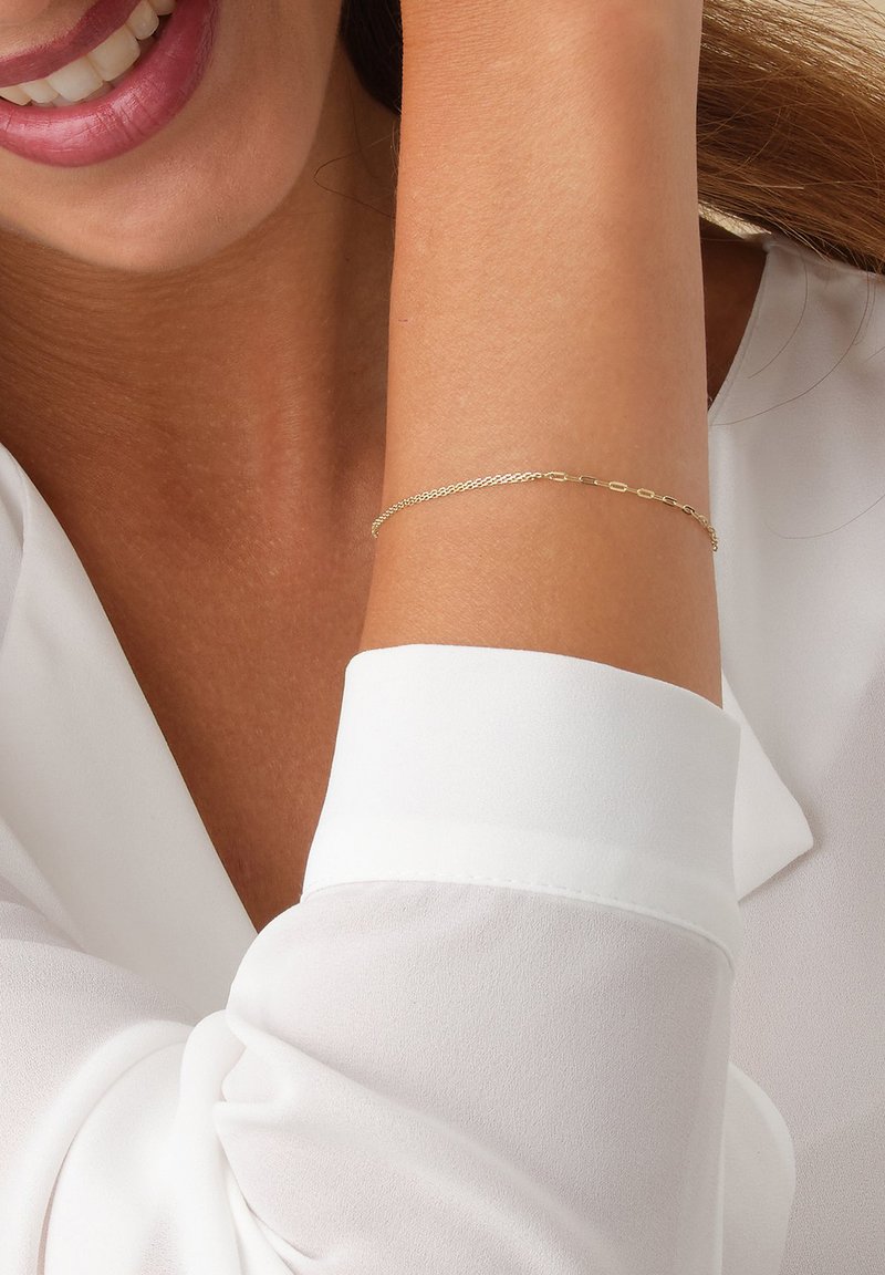 Lächelnde Person, die ein zartes goldfarbenes Armband am Handgelenk und eine weiße Bluse mit langen Ärmeln trägt, deren Manschette sichtbar ist.