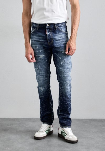Sötétkék farmernadrág, elhasználódott részletekkel és fakulással, egyenes fazonnal és zsebekkel, fehér sneaker cipőkkel, zöld csíkokkal párosítva.