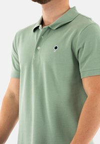 Camisa polo verde de algodón texturizado, con collar, dos botones y un pequeño logo de un árbol en azul marino en el lado izquierdo.