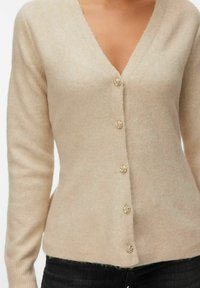 Femme portant un cardigan beige à col en V avec des boutons dorés en forme de rose, associé à un jean foncé, sur un fond blanc.