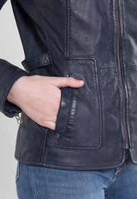 Mörkblå läderjacka med dragkedja, sidofickor och en figurnära design. Har en slät textur och kontrasterande sömmar.