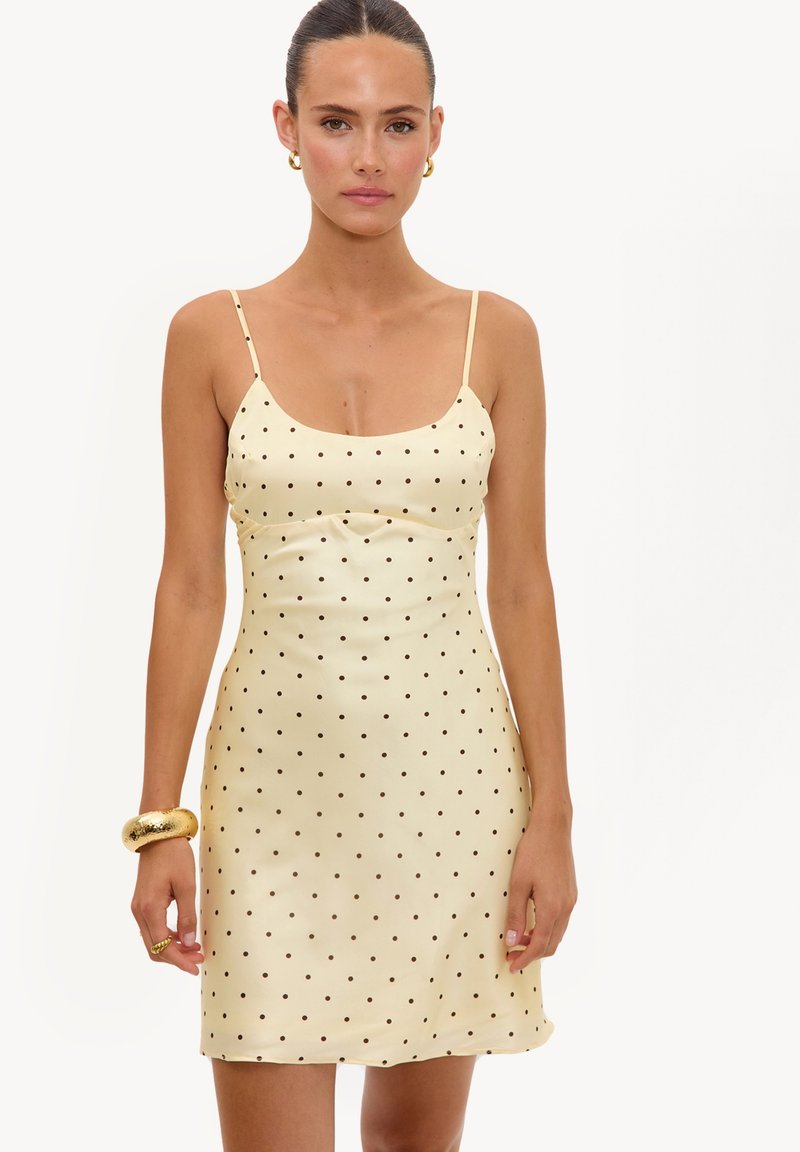 Femme portant une robe nuisette crème à pois noirs et fines bretelles, accessoirisée de boucles d’oreilles créoles dorées et d’un bracelet large.
