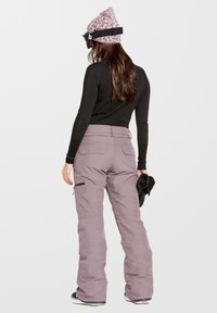 Pantalones de esquí morados con un diseño de pierna ancha, que cuentan con cremalleras laterales, bolsillos y una camiseta de manga larga negra texturizada. Sosteniendo guantes negros.