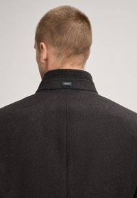 Mann mit kurzen Haaren, der von hinten zu sehen ist, trägt einen dunklen strukturierten Mantel mit einem sichtbaren "JOOP!"-Label am Kragen gegen einen schlichten Hintergrund.