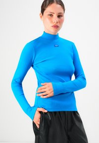 Kvinne iført en lys blå langermet turtleneck med "HUGO"-merke og svarte skinnbukser, poserer med en hånd i lommen og nøytral uttrykk.
