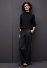 Un pullover à col roulé noir associé à un pantalon large gris foncé avec une bande latérale, complété par des chaussures noires à bouts pointus.