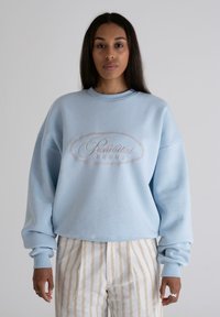 Sudadera azul claro con cuello redondo y mangas largas, que presenta un texto bordado "Prohibido" en un diseño ovalado decorativo.
