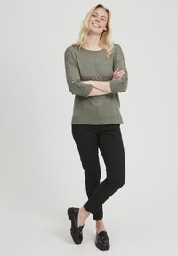 Olīvu zaļš krekls ar trim ceturtdaļām, pogu akcenti uz piedurknēm, kopā ar melnām šaurām biksēm un melniem mokasīniem.