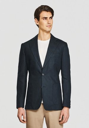 Mężczyzna w ciemnogranatowej marynarce, białej koszuli i beżowych spodniach, patrzący w bok na jednolitym jasnym tle.