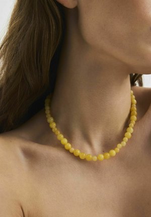 Donna che indossa una collana di perline rotonde gialle a vista ravvicinata, capelli castani lunghi dietro la spalla, collo e clavicola visibili.