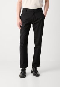 Pantaloni neri con una superficie liscia, design a gamba dritta e chiusura con bottone. Indossati con scarpe nere e una camicia beige.