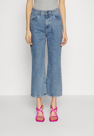 Moitié inférieure d'une personne portant un jean large court bleu clair et des sandales à talons hauts à brides rose vif sur un fond uni.