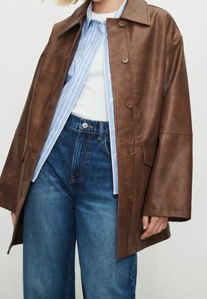 Frau trägt braune Lederjacke mit Knöpfen über einem blau-weiß gestreiften Hemd, darunter ein weißes Oberteil, das in blaue Jeans gesteckt ist.