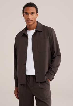 Jeune homme portant une veste-chemise marron foncé boutonnée sur un t-shirt blanc et un pantalon marron foncé assorti sur un fond clair.