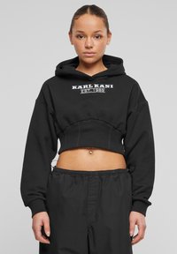 Must have lipsitudega kapuutsiga must lühikene sviiter, ribakujulise äärega ja valge "KARL KANI EST. 1989" logoga ees. Sobitatud mustade pükste juurde.