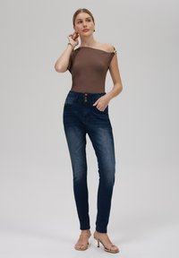 Brun off-shoulder bodysuit med gullaccenter, høy midje mørke denim skinny jeans med frontknapper og en glatt tekstur.