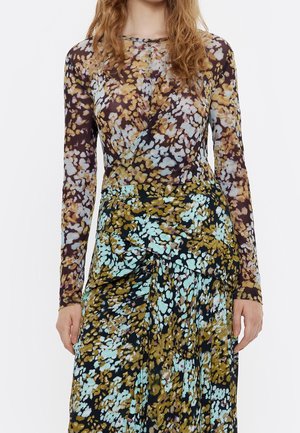 Femme portant une robe à manches longues avec un tissu superposé et un motif floral en marron, bleu et moutarde sur fond blanc.