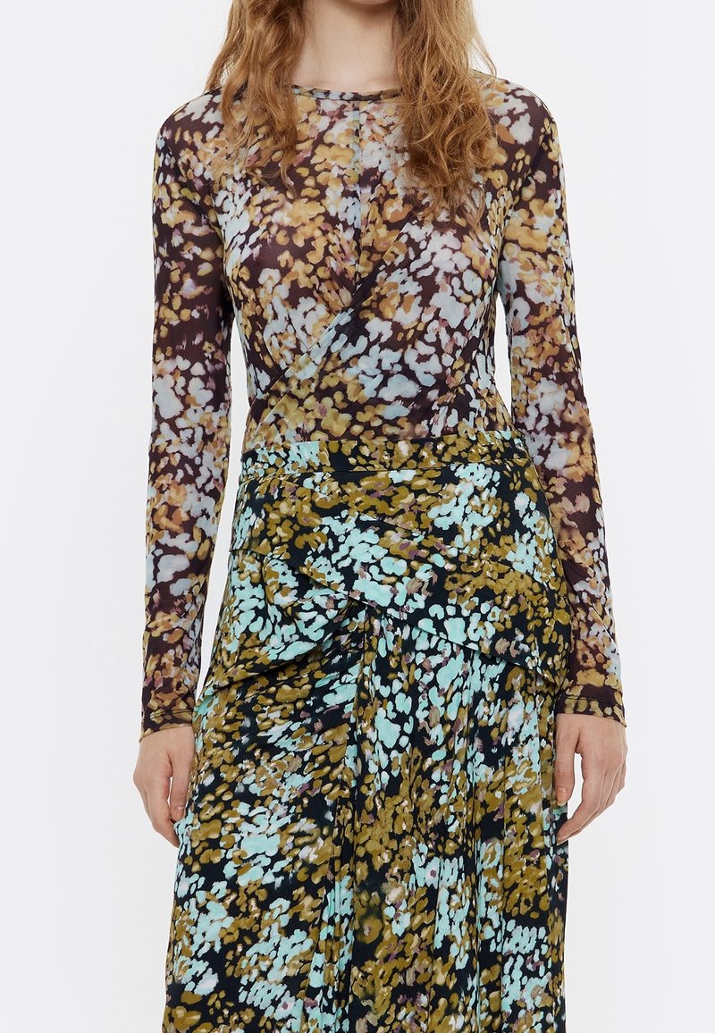 Femme portant une robe à manches longues avec un tissu superposé et un motif floral en marron, bleu et moutarde sur fond blanc.