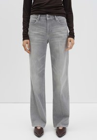 Persoon in lichtgrijze wijde jeans, zwart langmouwtop en donkere spitse schoenen, staande tegen een effen witte achtergrond.