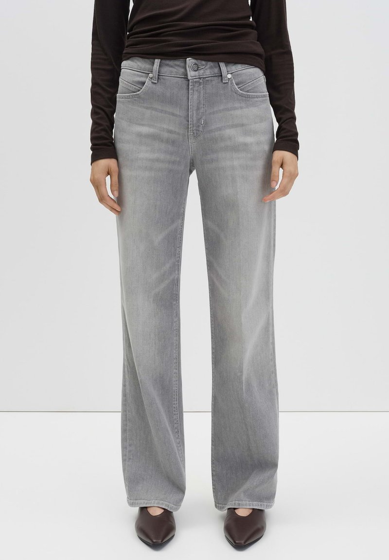 Persoon in lichtgrijze wijde jeans, zwart langmouwtop en donkere spitse schoenen, staande tegen een effen witte achtergrond.