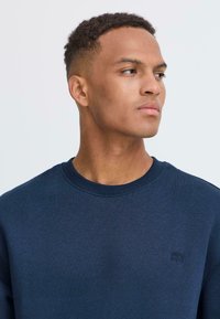 Mornarska majica s crew izrezom in rebrastimi manšeti ter spodnjim robom. Ima majhen, tonalen logotip na prsih. Material se zdi mehak in teksturiran.