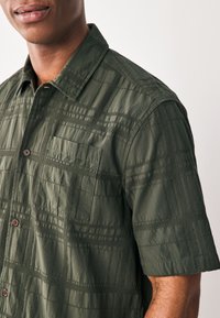 Dunkelgrünes T-Shirt mit kurzen Ärmeln, das ein strukturiertes Karomuster, eine Brusttasche und braune Knöpfe aufweist. Lässige Passform mit Kragen.