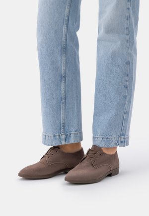 Jasnoniebieskie dżinsy w połączeniu z brązowymi zamszowymi oksfordami na osobie stojącej na białym tle.