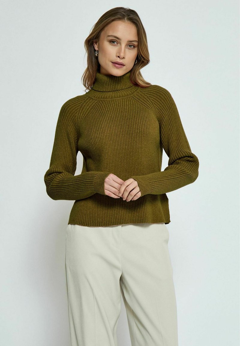 Temno olivno rebrasto turtleneck pulover z dolgimi rokavi, ki ima prilegajoče telo in sproščen rob, v kombinaciji z svetlo obarvanimi hlačami.