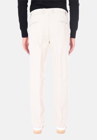 Persona che indossa pantaloni dritti color crema con tasche posteriori abbottonate, maglietta nera a maniche lunghe e scarpe bianche, in piedi contro uno sfondo bianco.