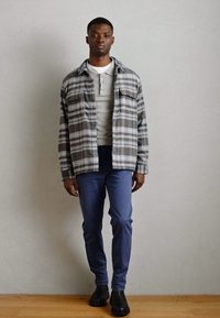 Karierte Flanellhemd in Grau mit weißen Streifen, mit Knopfleiste, graues Henley darunter, blaue Slim-Fit-Hosen und schwarze Stiefeletten.