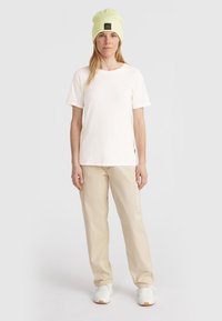 Witte t-shirt met korte mouwen, beige cargo broek en een lichtgroene beanie met een zwart logo patch. Model staat tegen een neutrale achtergrond.