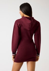 Abito felpa bordeaux con maniche lunghe, cappuccio profondo e dettagli a strisce sui polsini. Texture morbida, design aderente e lunghezza a metà coscia.