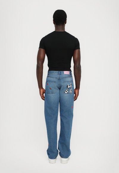 Jeans in denim blu con una vestibilità ampia, caratterizzati da un design di un cane ricamato sulla tasca posteriore e dettagli a forma di cuore. Indossati con una maglietta nera.