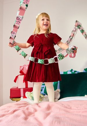 Fiatal lány piros ruhában, aki egy színes papírlánc girlandot tart, egy rózsaszín ágyon áll, mellette becsomagolt ajándékok és plüssjátékok vannak.