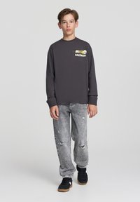 Maglietta grigia a maniche lunghe con grafica Pokémon sul petto, abbinata a jeans chiari strappati e sneaker nere.