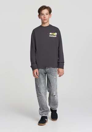 Maglietta grigia a maniche lunghe con grafica Pokémon sul petto, abbinata a jeans chiari strappati e sneaker nere.