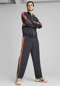 Námornícky modrá tepláková súprava s geometrickým vzorom a oranžovými pruhmi po bokoch. Bunda má predný zips; nohavice majú elastický pás.